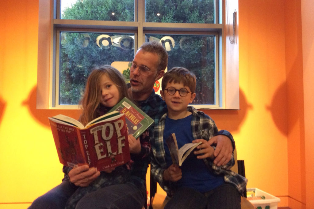 Image description: Andrew Dertien sits with his two children by the window, and appears to be reading aloud from a kids book. Each child holds a book as well. 