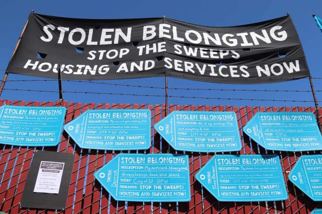 A banner reading "Stolen Belonging" is set up over placards shaped like tags describing the belongings stolen from homeless San Franciscans