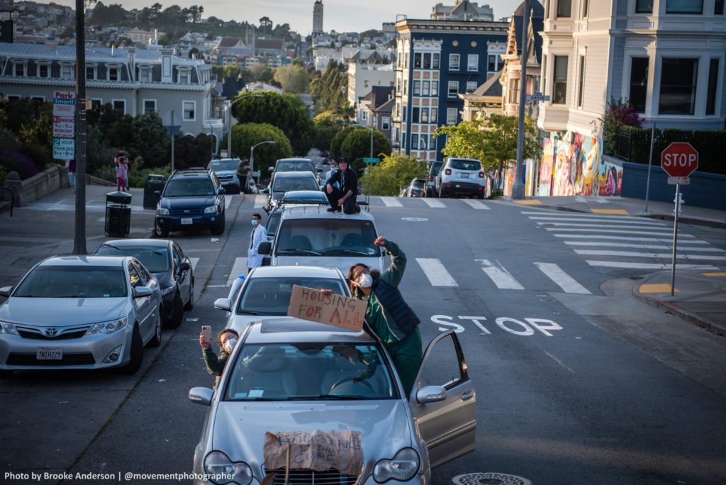 A line of vehicles stretches down a San Francisco street. People wearing face masks lean out the windows. A sign reads "Housing for all".