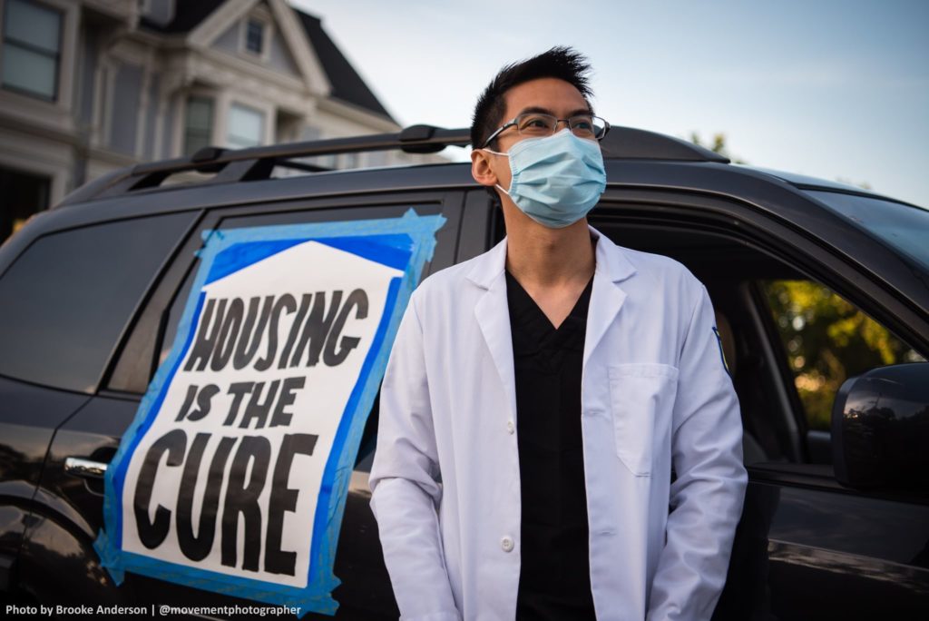 A person in a doctor's coat and face mask is in the foreground, behind them a vehicle with a sign taped to it that reads "HOUSING IS THE CURE"