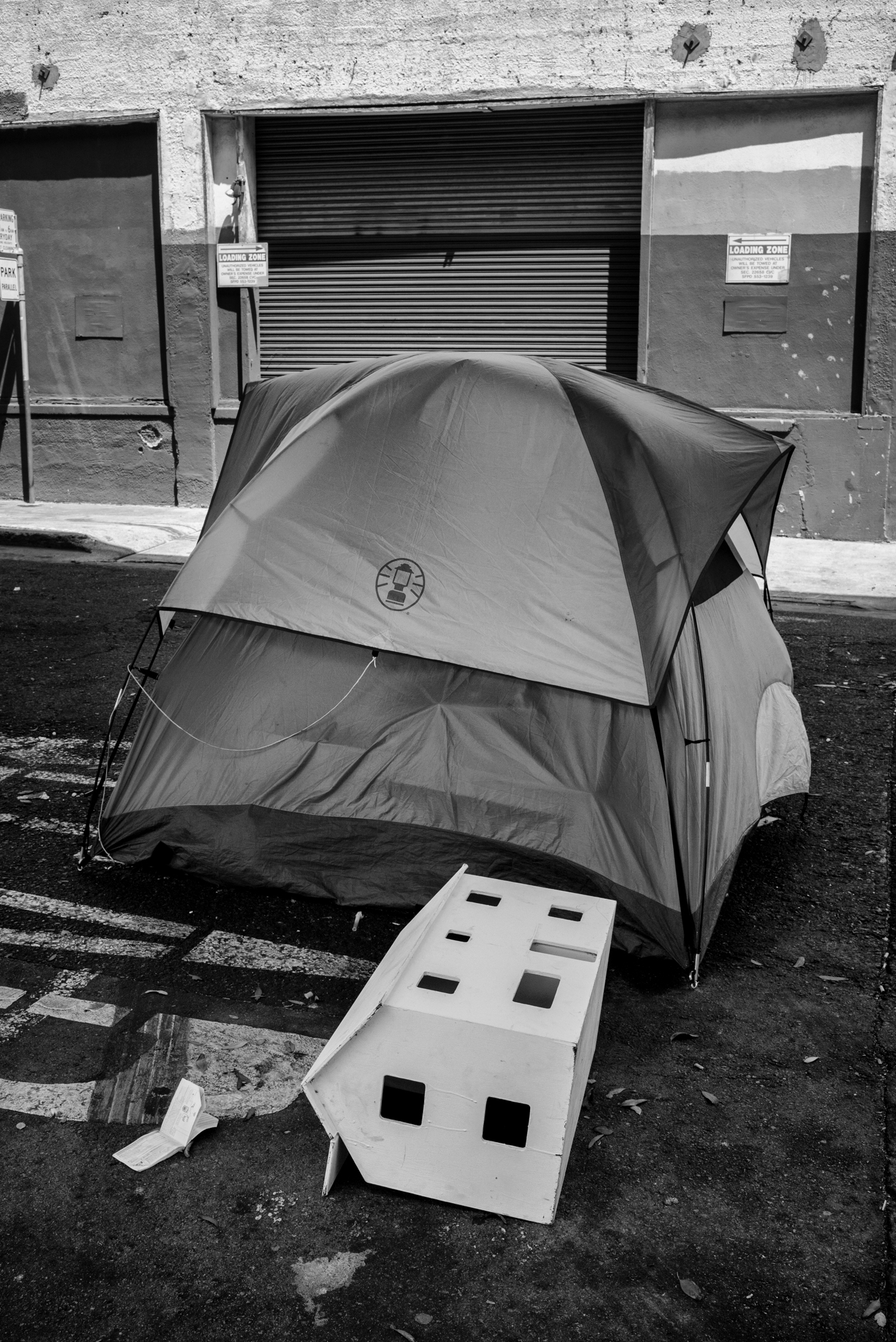 A tent is in the center of the frame. In front of it is what looks like a white dollhouse, laying flat on the ground. The image is in Black and White