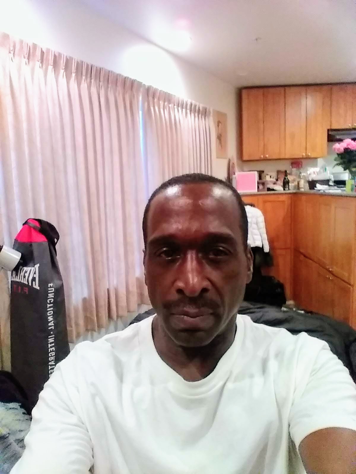 Selfie of a man in a white shirt in a room with bright curtains and wood cabinets in the background.