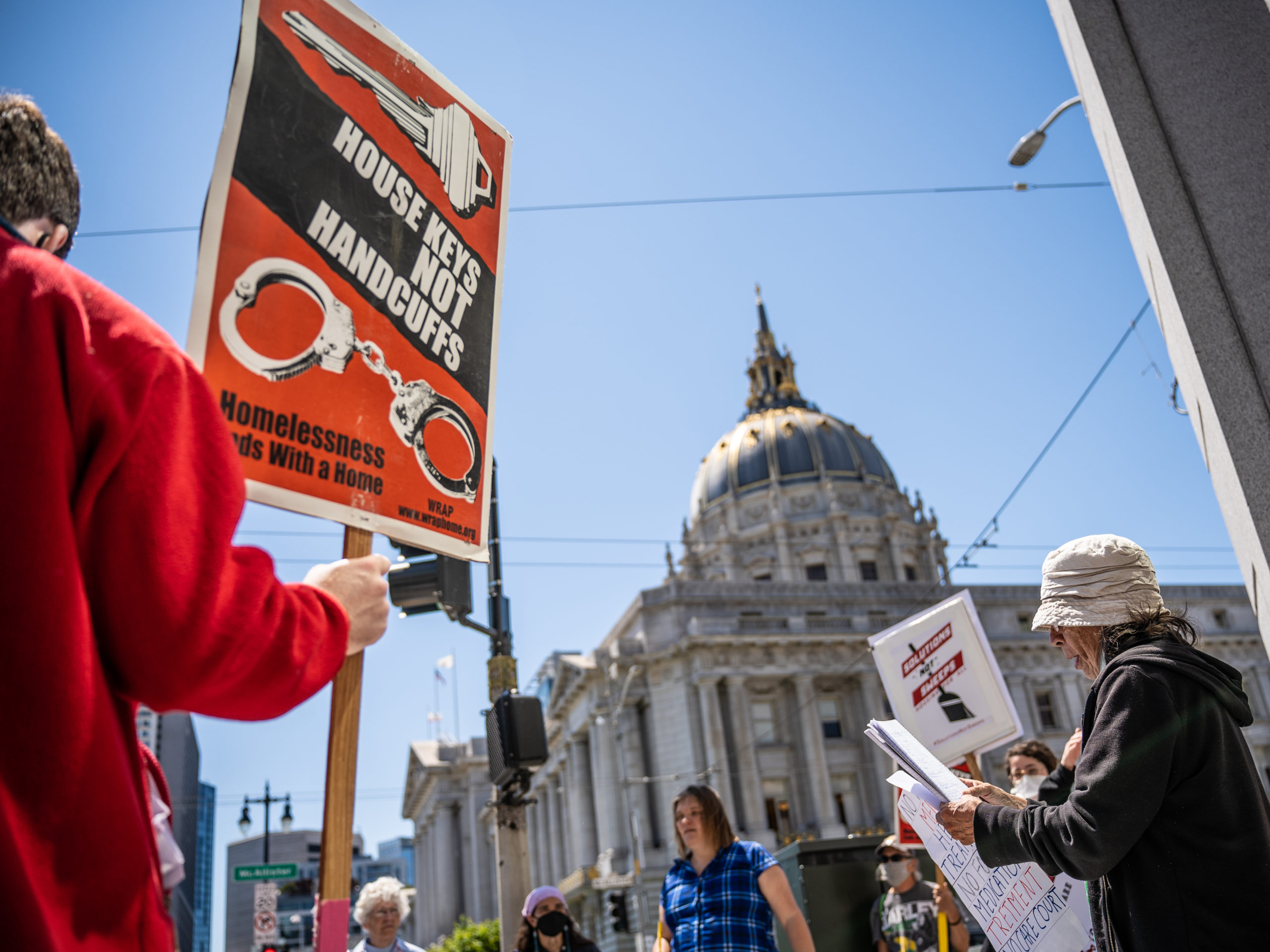 In the foreground we see a figure holding a sign that reads "Housekeys not Handcuffs", and a crowd is gathered. In the background San Francisco City Hall seems to loom.
