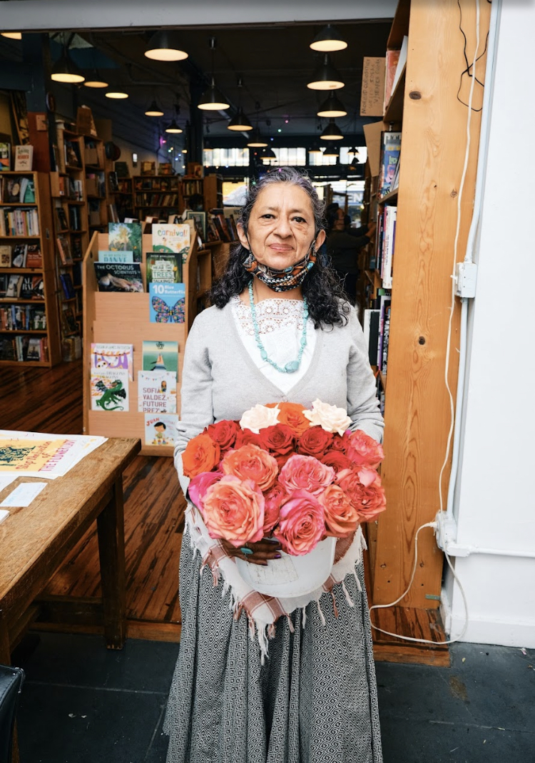Denhi the Flower Lady holding a pink bouquet, standing in a bookstore and smiling gently