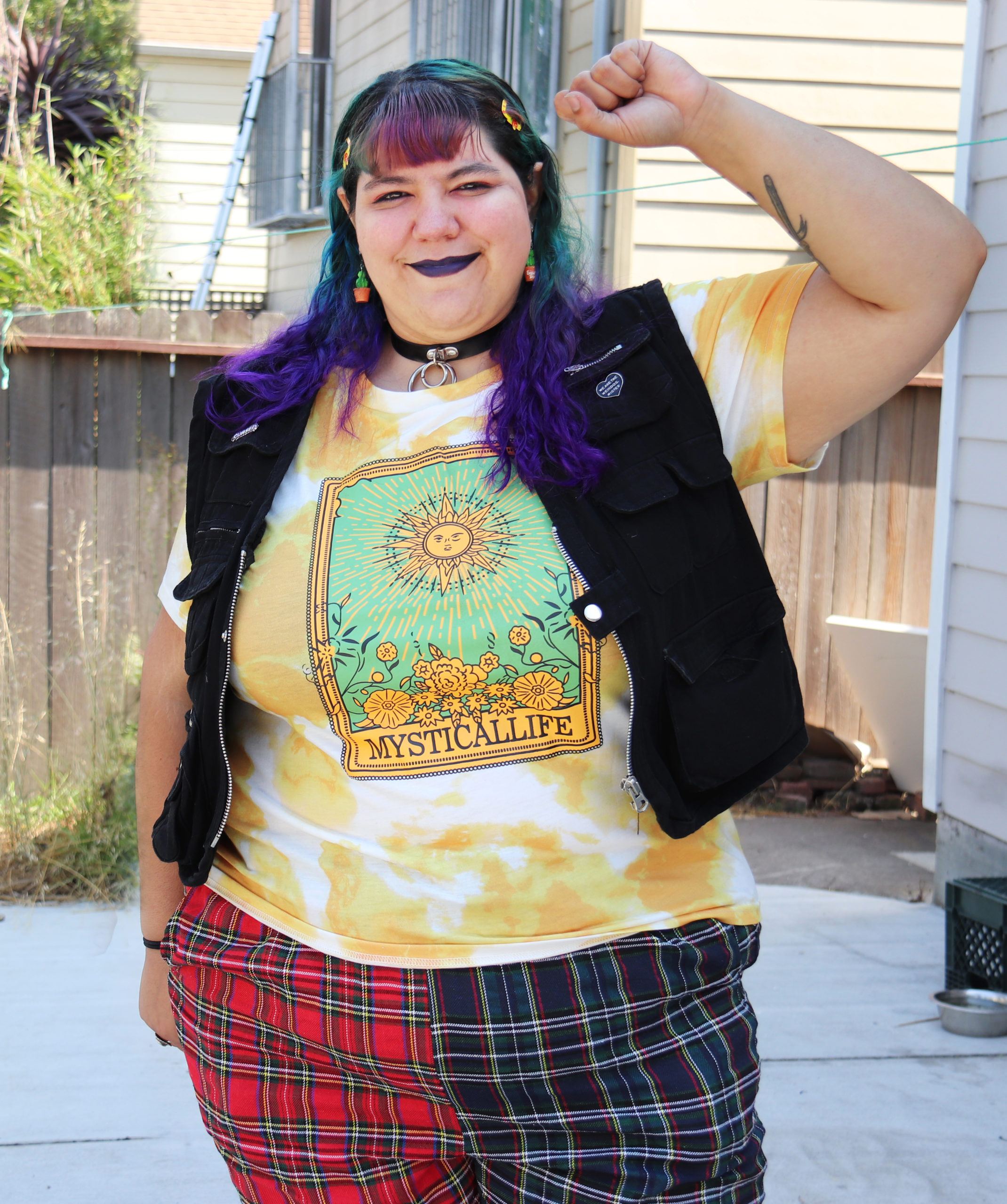 A woman with colorfully dyed hair raises her clenched fist and smiles at the camera. She is wear a graphic T-shirt, plaid pants, and a black vest.
